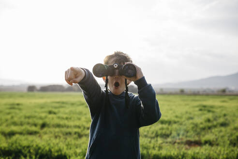 Surprised boy pointing while looking through binoculars in field during sunny day - JRFF05127