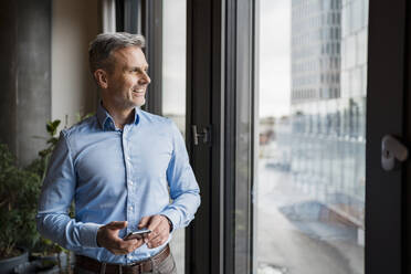 Smiling businessman looking through window while holding mobile phone at work place - DIGF15109