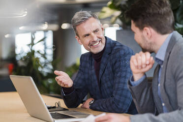 Smiling mature businessman gesturing while discussing over laptop with colleague in office - DIGF15146