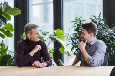 Smiling business people looking at each other while sitting at table in office - DIGF15159