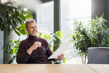 Lächelnder männlicher Unternehmer mit Brille und Papier träumt am Arbeitsplatz - DIGF15162