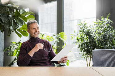 Smiling male entrepreneur with eyeglasses and paper day dreaming at work place - DIGF15162