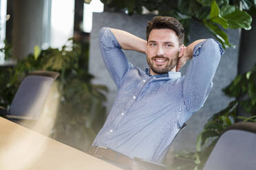 Smiling mid adult businessman with hands behind head relaxing at work place - DIGF15167