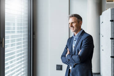 Smiling mature businessman with arms crossed day dreaming in office - DIGF15180