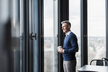 Smiling mature man holding coffee cup while looking through window at office - DIGF15207