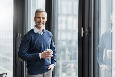 Lächelnde reife männliche Fachkraft mit Kaffeetasse stehend am Fenster im Büro - DIGF15209