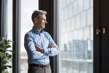 Smiling businessman with arms crossed day dreaming while standing at work place - DIGF15210