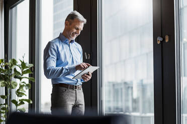 Smiling mature businessman working on digital tablet by window at work place - DIGF15211