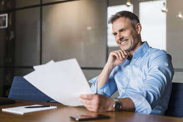 Smiling male professional with hand on chin examining paper documents in office - DIGF15214