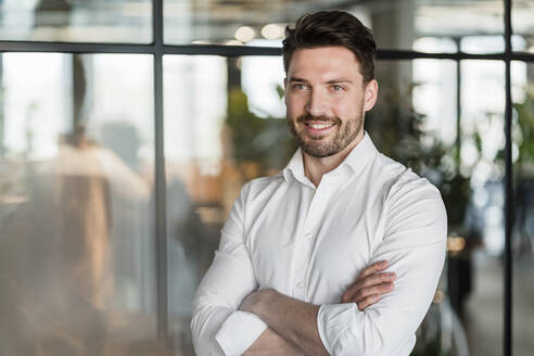 Lächelnde männliche Fachkraft mit verschränkten Armen, stehend an einem Glas im Büro - DIGF15227