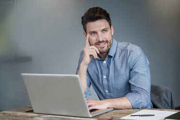 Nachdenklich lächelnder Geschäftsmann mit Laptop über Tisch am Arbeitsplatz - DIGF15241