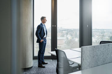 Mature businessman with hands in pockets looking through window in office - DIGF15249