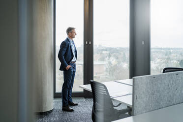 Reifer Geschäftsmann mit Händen in den Taschen schaut durch ein Fenster in einem Büro - DIGF15249