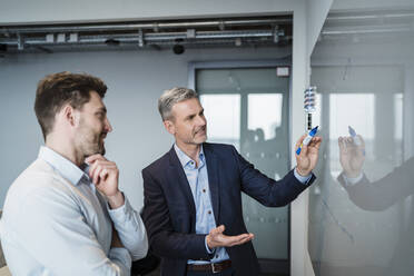 Reifer Geschäftsmann, der mit einem männlichen Kollegen im Büro über eine Grafik am Whiteboard diskutiert - DIGF15259