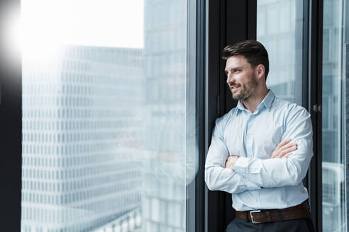 Lächelnder männlicher Unternehmer mit verschränkten Armen, der durch ein Fenster im Büro schaut - DIGF15277