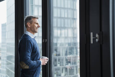 Smiling mature businessman with coffee cup looking through window at office - DIGF15283