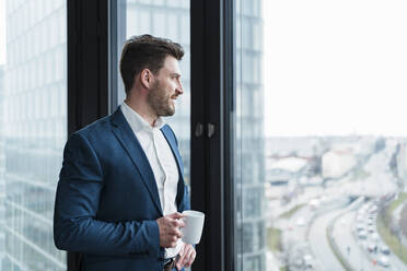 Businessman with coffee cup day dreaming by window at work place - DIGF15285