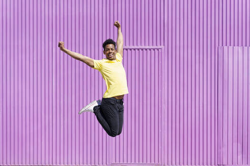 Carefree man jumping with arms raised by corrugated iron - JSMF02112