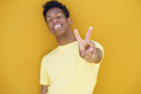 Smiling man showing peace gesture in front of yellow wall - JSMF02117