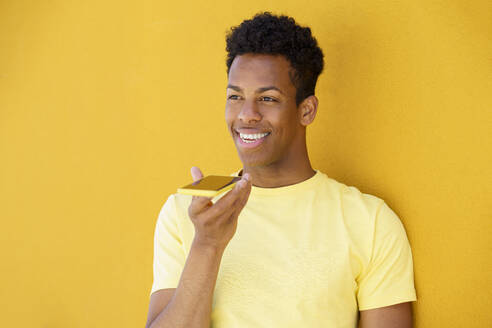 Smiling man making voice note through smart phone in front of yellow wall - JSMF02119
