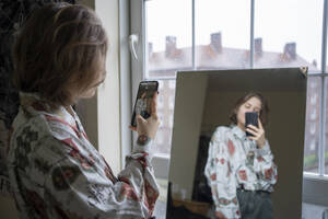 Junge Frau, die ihr Spiegelbild mit ihrem Smartphone zu Hause fotografiert - FBAF01790