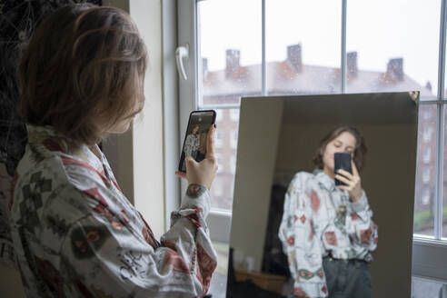 Young woman photographing reflection in mirror through smart phone at home - FBAF01790