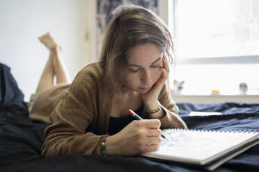 Hipster woman drawing on book while lying on bed at home - FBAF01795