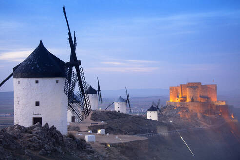 Spanien, Provinz Toledo, Consuegra, Historische Windmühlen in der Abenddämmerung mit der Burg von La Muela im Hintergrund - DSGF02412