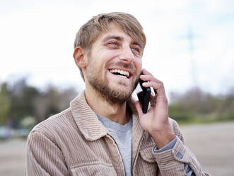 Happy bearded man looking away while talking on smart phone - FMKF07078