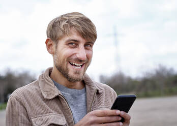 Smiling man with blond hair holding smart phone - FMKF07079