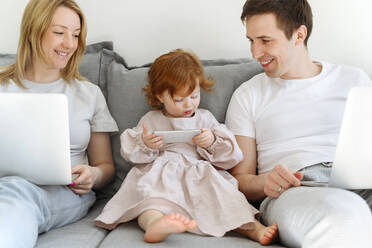 Smiling male and female entrepreneurs looking at daughter using mobile phone on sofa at home - VYF00543