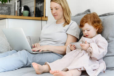 Girl using smart phone sitting by mother working on laptop at home - VYF00544