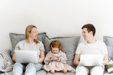 Smiling male and female entrepreneurs with laptop looking at each other while sitting by daughter on sofa in living room - VYF00545