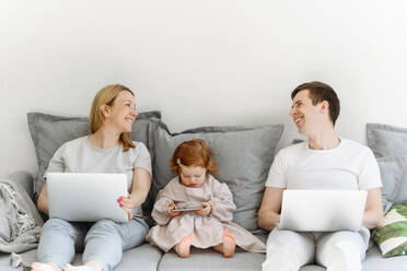 Happy male and female entrepreneurs with laptop smiling while sitting by daughter on sofa at home - VYF00546