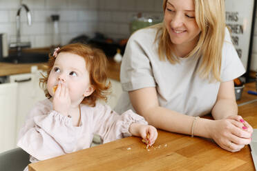 Smiling woman looking at daughter eating at dining table in kitchen - VYF00550