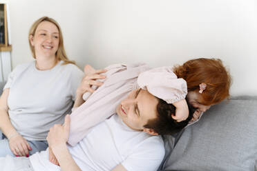 Lächelnde schwangere Frau, die ihre Tochter betrachtet, die mit ihrem Vater auf dem Sofa zu Hause spielt - VYF00554