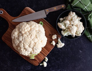 Blumenkohl und Röschen auf einem Schneidebrett und in einer Schüssel auf einer schwarzen Arbeitsplatte. - CAVF93850