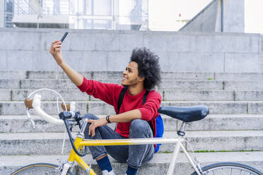 Junger Mann mit Afro-Haar sitzt und macht ein Selfie neben seinem alten Fahrrad - CAVF93857