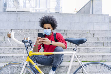 Young man with afro hair sitting sending a message next to his old bicycle - CAVF93859