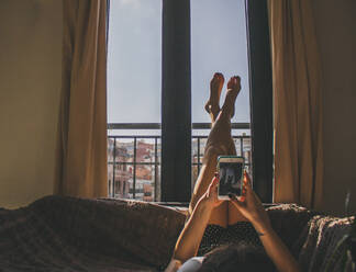 Junge Frau auf dem Sofa liegend mit hochgelegten Füßen beim Fotografieren - CAVF93873