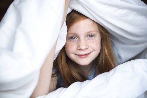 Young red haired girl peeking out from under a white comforter. - CAVF93887
