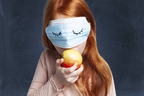 Girl eating apple while covering eyes with protective face mask - GAF00151