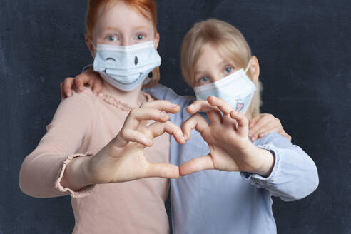 Sisters making heart shape with hands wearing face mask against black background - GAF00152