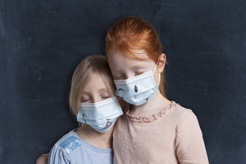 Sisters with eyes closed wearing protective face mask against black background - GAF00155