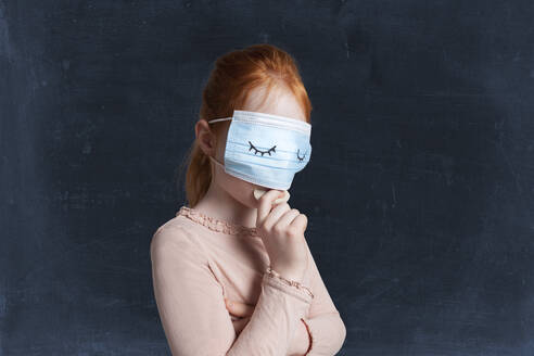 Girl eating biscuit while wearing protective face mask against black background - GAF00157
