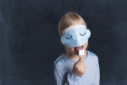 Mädchen mit Gesichtsschutzmaske leckt Lutscher vor schwarzem Hintergrund - GAF00159