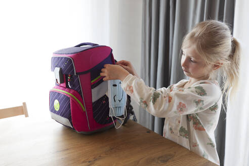 Girl tying face mask on backpack at home - GAF00176