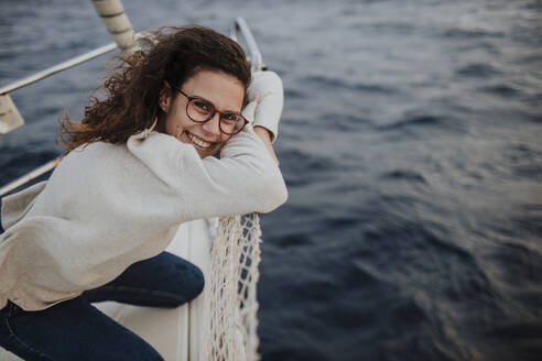 Glückliche Frau mit Brille, die sich im Urlaub auf einem Boot an ein Netz lehnt - GMLF01175