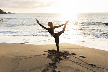 Woman stretching while practicing yoga by water's edge at beach - JPTF00737