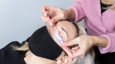 Professional beautician applying artificial eyelashes on young female client wearing protective face mask in modern beauty studio - ADSF22697